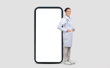 Steteskoplu beyaz laboratuar önlüklü gülümseyen Avrupalı kadın doktor, teletıp ve modern sağlık hizmetlerini sembolize eden büyük boy akıllı telefonun yanında duruyor.