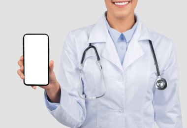 Laboratuvar önlüklü gülümseyen Avrupalı bir bayan doktor, gri stüdyo arka planında izole edilmiş, tıbbi uygulamalar veya tele-sağlık hizmetleri için hazır, boş ekranlı bir akıllı telefon sunar.