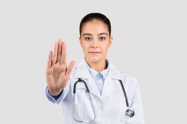 Steteskoplu ciddi genç Avrupalı doktor elleriyle işaret etmeyi keserek, sağlık güvenlik önlemleri ve sınırlarının önemini vurgulayarak, gri stüdyo arka planında izole edilmiş,