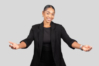 Mutlu Afro-Amerikan iş kadınlarını davet etmek, açık elleri ile hoş geldin ya da sunum pozu veren, güler yüzlü, profesyonel siyah takım elbiseli, gri arka planda, stüdyo
