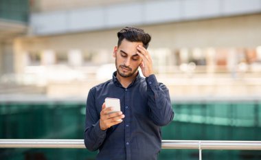 Endişeli Ortadoğulu genç işadamı akıllı telefonu kontrol ediyor, telefonundaki haberlerden rahatsız oluyor, stres belirtileri gösteriyor, açık alanda mesajlaşıyor.
