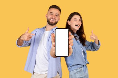 Neşeli erkek ve kadın başparmaklarını kaldırıyor ve boş akıllı telefon ekranına işaret ediyor. Uygulama promosyonu için ideal, güneşli sarı arka plana karşı.