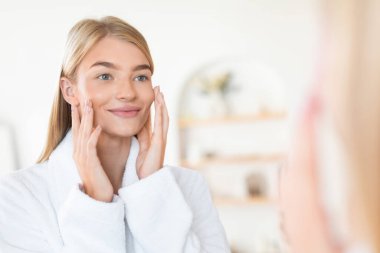 Sarışın kadın yüzlerine dokunup aynaya bakarken gülümsüyor modern banyo ortamında cilt bakımı sonrası mükemmel yumuşak teninin tadını çıkarıyor. Seçici odak, boş alan