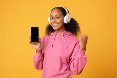 Müzik uygulaması. Mutlu siyah genç kız, sarı stüdyo arka planında müzik dinlerken kulaklık takıp, el kol hareketi yaparak telefonun boş olduğunu gösteriyor. Uygulama reklamı için model