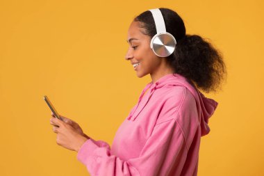 Smartphone kullanan, Kulaklık takan, Çevrimiçi Müzik Dinleyen Sarı Stüdyo Arkaplanı Üzerindeki Siyah Genç Kızın Yan Görünümü. Bayanın Favori Şarkısı 'nın tadını çıkarması. Müzikal Uygulama