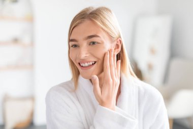 Parlak sarışın bayan yüzüne dokunuyor güzellik tedaviden sonra yumuşak tenini hissediyor, evdeki banyoya özgüven yayıyor. Cilt ürünleri ve güzellik bakım rutin konsepti
