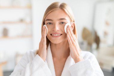 Bornozlu genç sarışın kadın yanaklarına pamuk yastık bastırıyor, kameraya gülümsüyor, sıcak güneşli banyoda cildini kucaklıyor. Tazelenen cilt tedavisi