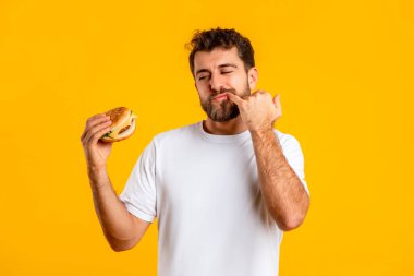 Suçlu Zevk. Elinde hamburger tutan, sağlıksız fast food yiyen, sarı stüdyo arka planında aç komik adamın portresi. Heyecanlı aç adam çizburgerin parmaklarını yalamak istiyor. Lezzetli.