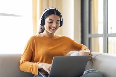 Neşeli Hintli kadın evde dizüstü bilgisayarıyla çalışırken kulaklıkla müzik dinlemekten zevk alıyor, gülümseyen kadın elinde bir fincan kahve, güneşli bir oturma odasında kanepede uzanıyor, yakınlaşıyor.