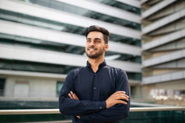 Neşeli Arap yönetici iş adamı, başarılı iş kariyeriyle gurur duyarak, gülümseyerek bir kenara bakarken, modern ofis binalarının dışında kendine güvenle silah doğrultuyor.