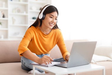 Gülümseyen genç Asyalı kadın evden ders çalışıyor, kanepede oturuyor, kablosuz kulaklık kullanıyor, dizüstü bilgisayara bakıyor, not alıyor, çevrimiçi derse katılıyor, eğitime katılıyor, Webinar 'ı kopyalıyor.