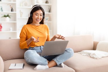 Pozitif, genç Asyalı bir kadın online ders alıyor, kanepeye oturuyor, bilgisayarlı dizüstü bilgisayar kullanıyor, evde kablosuz kulaklık takıyor, öğretmenle konuşuyor, kopyalama alanı kuruyor. E-eğitim, webinar, online okul