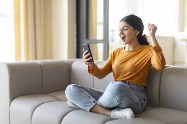 Evde başarıyı kutlayan akıllı telefonlu neşeli Hintli kadın cep telefonu ekranına bakarken muzaffer bir şekilde yumruğunu kaldırıyor, zaferi mi yoksa iyi haberi mi alkışlıyor?