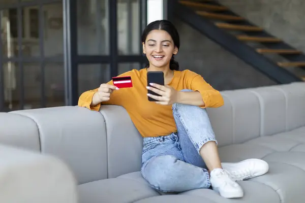 Relaxed young indian woman using credit card and mobile phone for online shopping, happy eastern female sitting on grey sofa in stylish home interior, making internet purchases, copy space