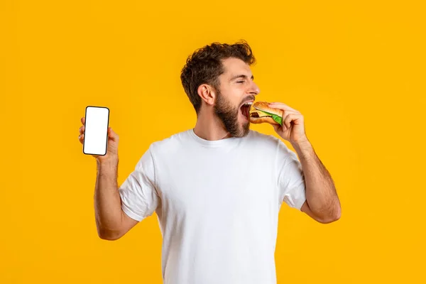 Hongerige Jongeman Met Hamburger Toont Smartphone Leeg Scherm Eten Gele — Stockfoto