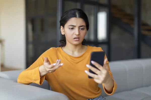 Spam Mailout. Annoyed young indian female looking at smartphone screen while sitting on couch at home, eastern woman feeling angry of spamming in electronic messages, reading bad sms, copy space