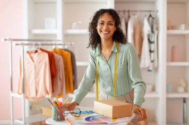 Neşeli Latin moda tasarımcısı elinde paketlenmiş ürün ve renk örnekleri, somutlaştırıcı yaratıcılık ve modern moda atölyesinde girişimci ruhu olan iyi ışıklandırılmış bir stüdyoda duruyor.