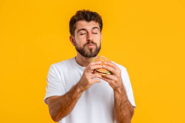 Abur cubur. Aç Avrupalı Genç Adam Lezzetli Burger Yiyor, Sarı Stüdyo Arkaplanına Karşı Ayakta Duruyor. Sağlıksız Rahatsızlık Yemeği 'nin tadını çıkaran adam. Dengesiz Beslenme ve Hile Yemekleri