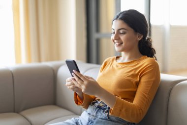Gülümseyen Hintli genç bir kadın evdeki kanepede dinlenirken akıllı telefonunu karıştırıyor, mutlu bir doğu kadını cep telefonu kullanıyor, mesajlaşıyor ya da internetten alışveriş yapıyor, rahat bir oturma odasında kanepede oturuyor.