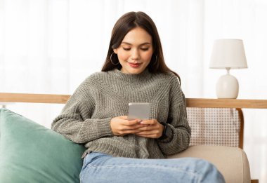 Evdeki kanepede dinlenen neşeli, çekici genç bir kadın, akıllı telefon kullanıyor ve gülümsüyor, arkadaşlarına mesaj atıyor, flört uygulamasında erkeklerle sohbet ediyor, internetten alışveriş yapıyor, fotokopi çekiyor.