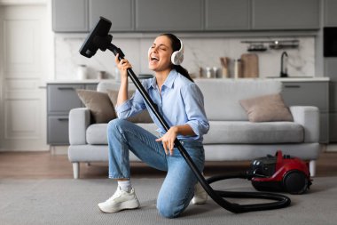 Enerjik genç bir kadın temizlik yaparken eğleniyor, elektrikli süpürgeyi gitar olarak kullanıyor, mutlu bir şekilde şarkı söylüyor ve modern bir oturma odasında kulaklık takıyor.