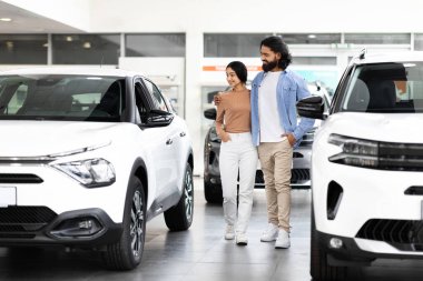 Neşeli genç doğu çifti, galeri otomobil salonunda yeni lüks arabaları kontrol ediyor. Mutlu milenyum eşleri sergi salonunda modern otomobili kucaklıyor ve seçiyor.