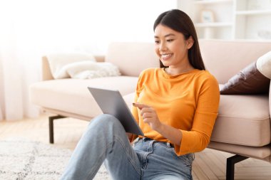 Uzun saçlı mutlu Asyalı kadın yerde oturuyor, dijital tablette haberler okuyor, online video içeriği izliyor, kopyalama alanı izliyor. Çinli kadın hafta sonunun keyfini çıkarıyor.