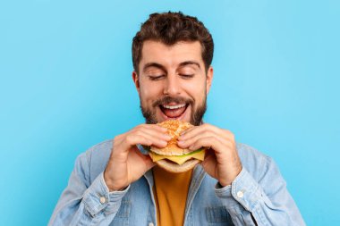 Abur cubur fazla yeme. Mutlu beyaz adam hamburger yiyor ve gülümsüyor, lezzetli sağlıksız atıştırmalıkların tadını çıkarıyor, mavi stüdyo arka planında dikiliyor, portre çekimi yapıyor. Hile yemek, beslenme ve erkek diyeti konsepti