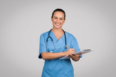 Gülümseyen beyaz kadın doktor, mavi üniformalı hemşire, panoya not alıyor ve gri stüdyo arka planında izole edilmiş kameraya gülümsüyor. Tıbbi muayene, sağlık, profesyonel yardım.