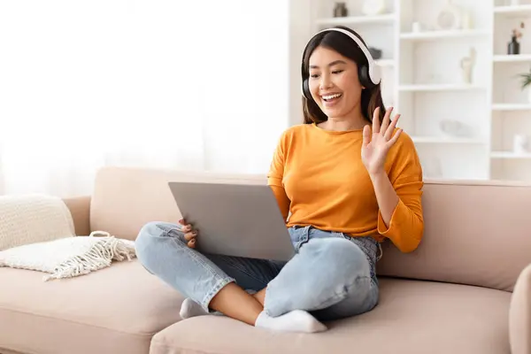 Positive millennial chinese lady have online meeting with family or friends, sit on couch with computer laptop on her lap, wave at pc screen and smiling, home interior, copy space. Telecommunication
