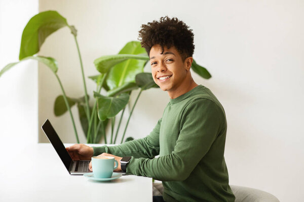 Cheerful stylish young black guy freelancer working from cozy cafe, sitting at desk, using laptop computer, drinking coffee in paper cup, smiling at camera, side view, copy space. Remote job