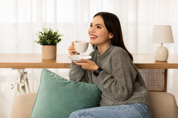 快乐的年轻女子一边喝着芬芳的咖啡 一边躺在沙发上 凝视着空旷的空间 微笑的黑发女士 喝点热饮料 休息时放松一下 — 图库照片