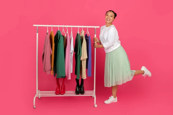 Smiling Asian lady standing beside garment rack filled with trendy clothes, happy cheerful korean shopaholic lady posing on pink studio background, enjoying fashion and seasonal slaes, copy space