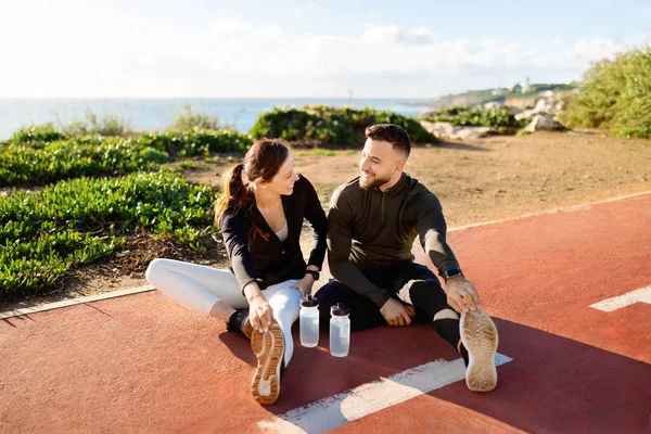 Joyful man and woman in sportswear sitting and stretching on running track with water bottles, sharing a moment of wellness and laughter