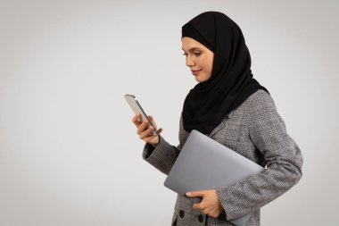 Tesettürlü profesyonel bir kadın bilgisayarını kapalı tutarken dikkatli bir şekilde akıllı telefonunu kullanıyor, iş kıyafetleri içinde sade bir arka plana karşı duruyor.