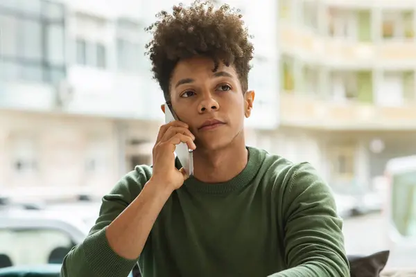 Closeup of handsome young black guy with curly hair sitting at table by window at cafe, have phone conversation with friend or lover, looking at copy space. Communication and technologies