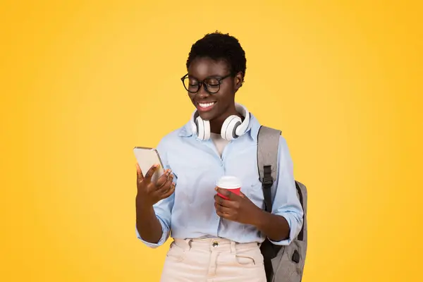 Glad black woman student in glasses, multitasking with coffee in hand and phone, isolated on yellow background. Study, busy lifestyle, chat in social networks, break and education