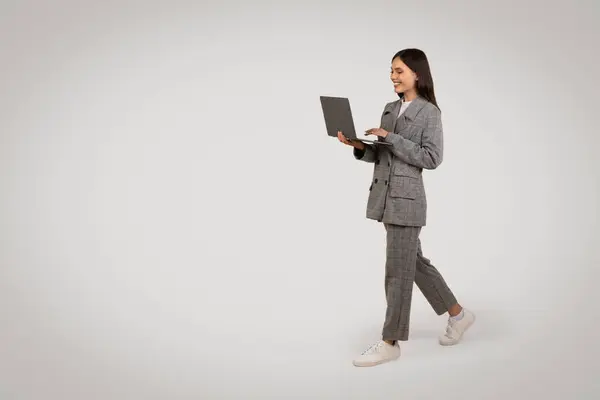 アクティブな若いプロの女性ウォーキングとノートパソコンを使用して カジュアルなスニーカーでドレスを着て 最小限のグレーの背景 無料スペースに対して — ストック写真