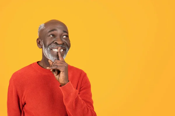 Cheerful senior african american man put finger to his lips in a shushing gesture, wearing a bright red sweater against orange studio background, suggesting silence. Secret, gossip, news