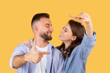 Neşeli çift parmaklarıyla çerçeve yapar, eğlencenin görüntüsünü yakalar, aynı zamanda neşeli sarı arka plana karşı gülümseme takınırlar.