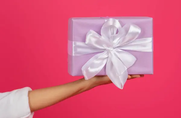 Female hand presenting beautifully wrapped gift box with white satin ribbon, unrecognizable lady offering sense of celebration and surprise, giving present, standing against vibrant pink background