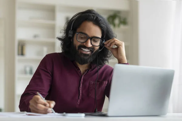 Happy indian man with headset taking notes and using laptop at home, smiling eastern male sitting at desk, engaged in cheerful conversation, exemplifying effective remote communication, closeup