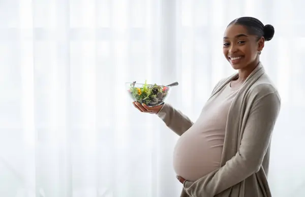 幸せな若い黒人女性は 彼女の大きな腫れを抱きしめ 自宅の窓の隣に立って新鮮な有機野菜サラダでボウルを保持することを期待しています 健康的な食事 妊娠中の女性のための栄養 — ストック写真