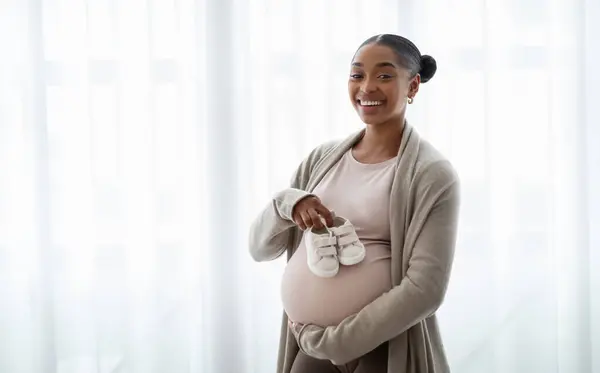 自宅の窓の隣に立っている妊婦の腹の胎児のための小さな靴 子供は期待する女性の腹の未生児のためのバラバラ コピースペース付きのパノラマ — ストック写真