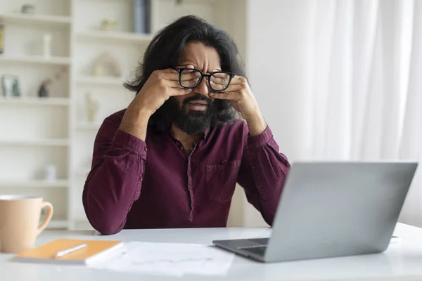 Tired indian man rubbing eyes, suffering from eyestrain during working on laptop, young eastern male freelancer feeling exhausted, sitting at desk at workspace in home office, free space