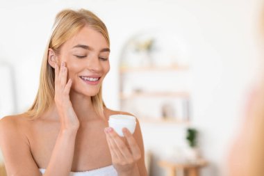 Genç Avrupalı sarışın kadın cildini kozmetiklerle şımartıyor, yüz kremi sürüyor, ev içi cilt bakımını kucaklıyor, sıhhatli bir yaşam sürüyor ve sıcak bir banyoda mutlu bir yaşam sürüyor. Boş alan