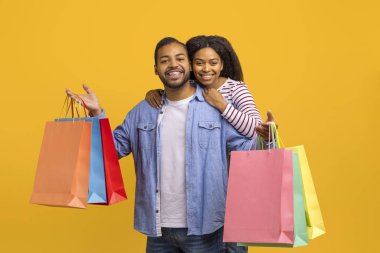 Sarı arka planda alışveriş torbaları olan neşeli siyah çift pozları, mutlu Afrika kökenli Amerikalı erkek ve kadın alışverişleri, sezonluk satışların keyfini çıkarması, kopyalama alanı.