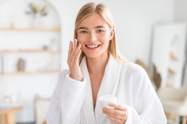 Yüzüne nemlendirici sürerken kameraya gülümseyen genç sarışın kadın, yüz bakımı ve açık banyo içinde cilt ayinlerinden zevk alıyor, bornozla kameraya gülümsüyor.