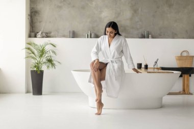 Beyaz bornoz giyen güzel Hintli bir kadın modern lüks banyoda küvette oturuyor, bacaklarına dokunuyor, epilasyon sonrası pürüzsüz ipek tenin tadını çıkarıyor veya tıraş oluyor, kopyalama uzayı.