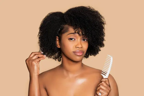 Young black woman appears sad as she touches her curly afro hair and holding white comb, standing against soft beige background